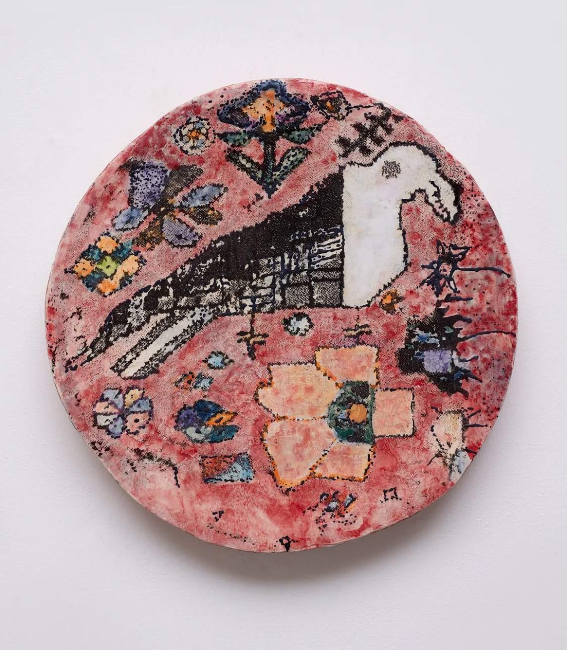 This flat stoneware disk is painted with touches of black, rust red, coral orange, and shades of purple and blue to resemble a woven rug. A white and black bird at the center is surrounded by flowers against the red background.