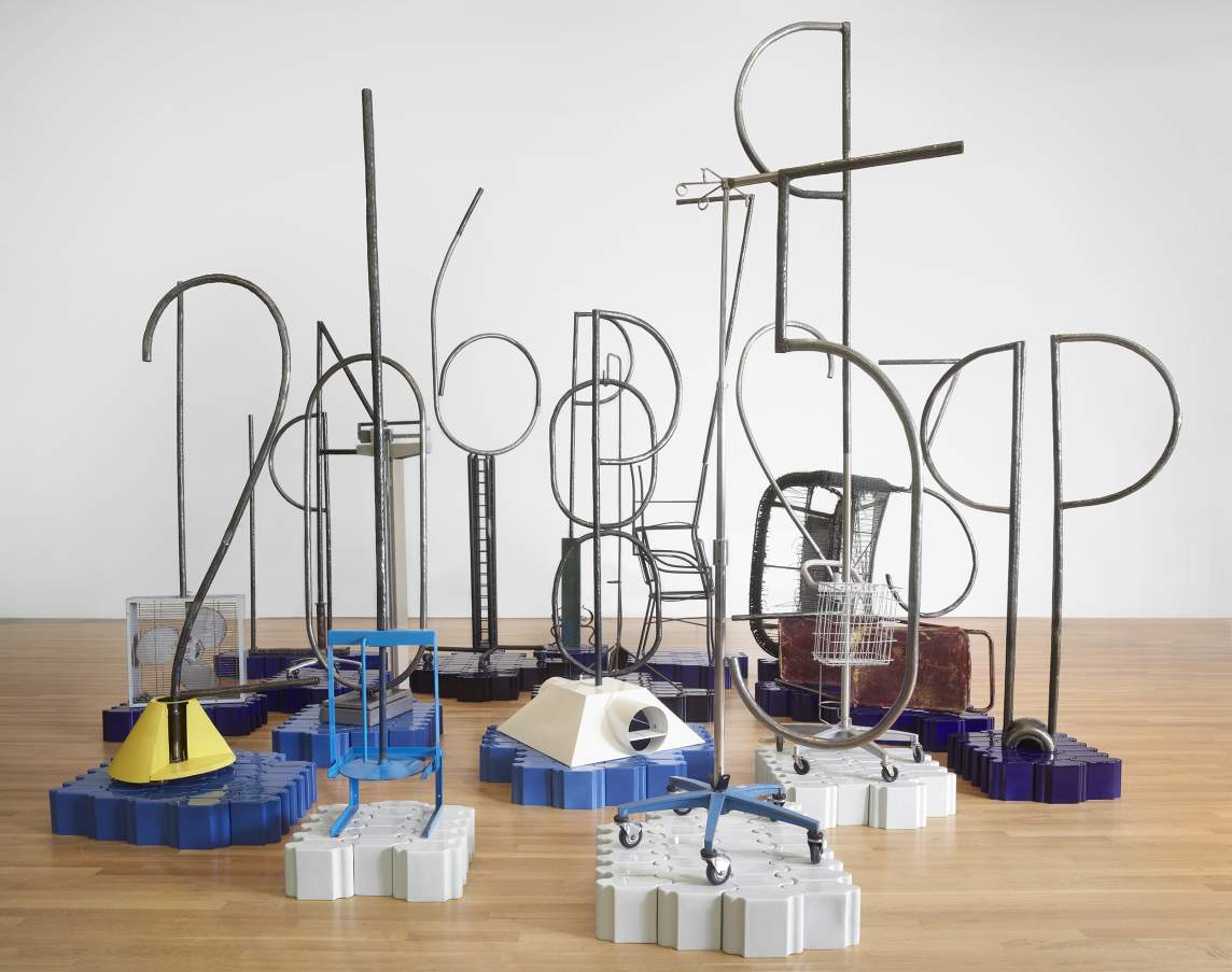 Metal rods formed into numbers are held up by wheeled stands or metal frames, except for two, which are braced in a mechanical beam scale and on a box fan respectively. Each digit and base are set on individual islands of tall, interlocking tiles in shades of blue, white, or purple on the gallery’s wood floor.