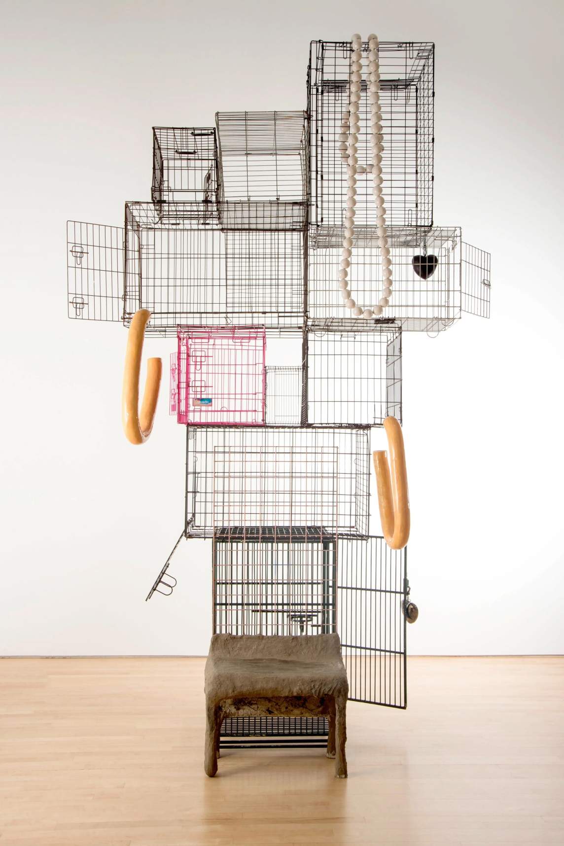 Five levels of thin-wired cages are piled behind a low, square seat. Each level gets wider except the topmost, which is nearly the same width as the fourth level, and some of the cage doors hang open. Most of the cages are black or metal, and one is hot pink. An oversized string of white beads hangs over one of the topmost cages. A black heart, like a pendant, hangs inside one cage nearby, and yellow hoops hang from lower levels.