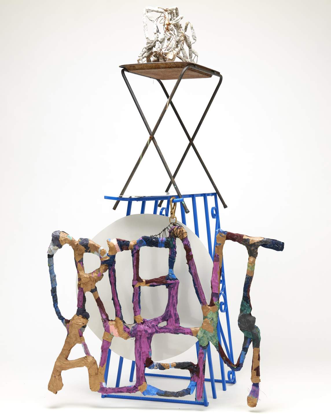 Untitled (Orientalism charm with TV tray) is a mixed media assemblage structure. At the base, sculptural words read “ORIENTALISM, in front of a white steel disc, and blue security bars. On top, a TV tray teeters, upon which rests a white sculptural rubble of tangled ceramic, concrete, and wire.