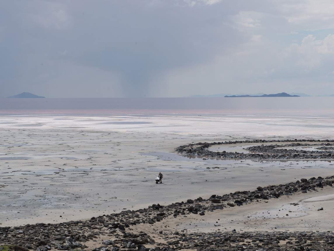 On a vast, tranquil salt flat adjacent to Robert Smithson's land art sculpture Spiral Jetty, the artist William Lamson gathers materials in a wheelbarrow, surrounded by subtle patterns of dry salt and distant mountains under a cloudy sky.
