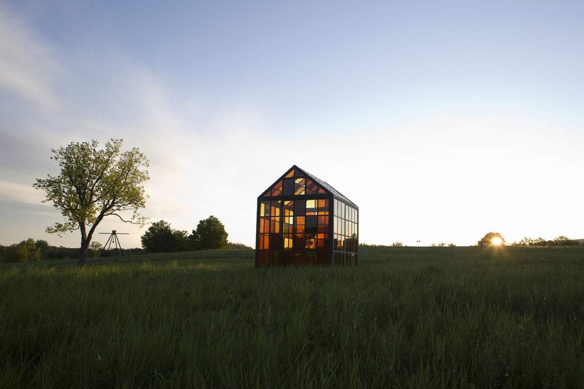 “Solarium” is a greenhouse-like sculpture set on a grassy hill at dusk, formed of gridded panes of yellow, brown, and honey colored glass. Tree saplings are visible through the windows.