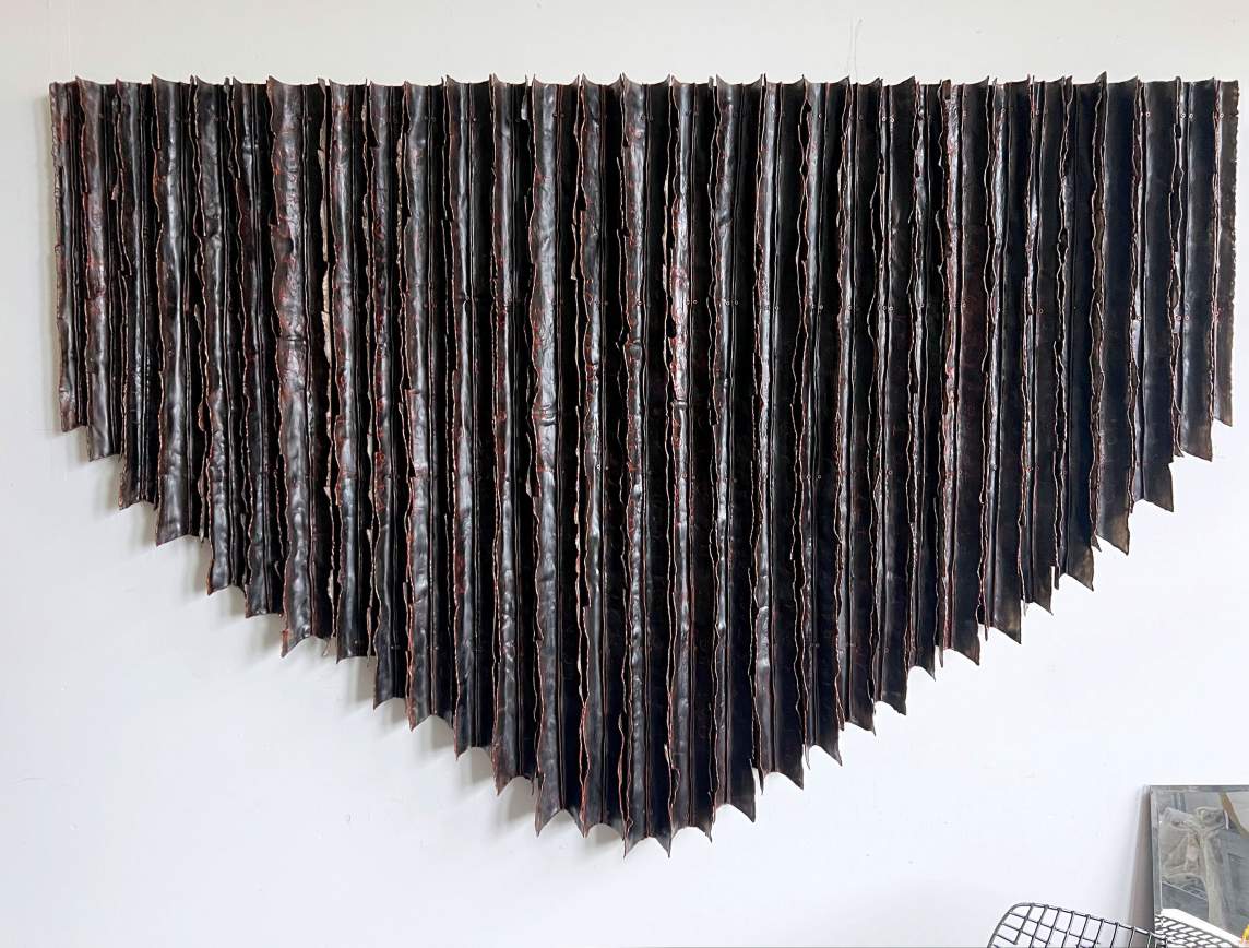 Thirty dark brown strips folded into long V shapes are connected to make a sculpture that hangs on a wall. The sculpture is straight across the top, and the lengths vary to make the bottom edge create a wide V. Light glinting off the surfaces suggest that the material has a sheen.