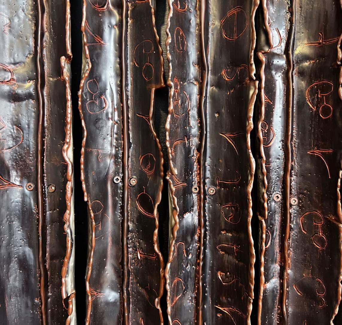 Close-up of four strips. Each is pleated down the center and the flaps created by the V-shaped folds are irregular along their edges. The material is dark brown, but the edges and characters from the Cherokee Syllabary along the flaps are copper colored. The strips are held in place with screws.