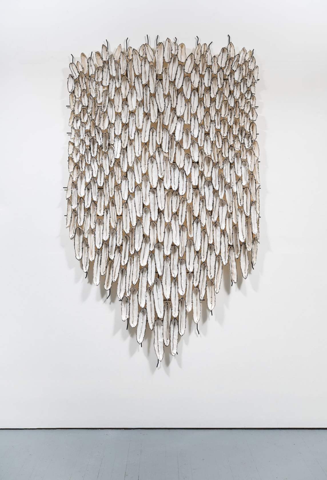 A few hundred tongue-like shapes overlap and hang like feathers or scales in a rough shield-like form on a white gallery wall. Each shape is white outlined in black and has a black line down the center.