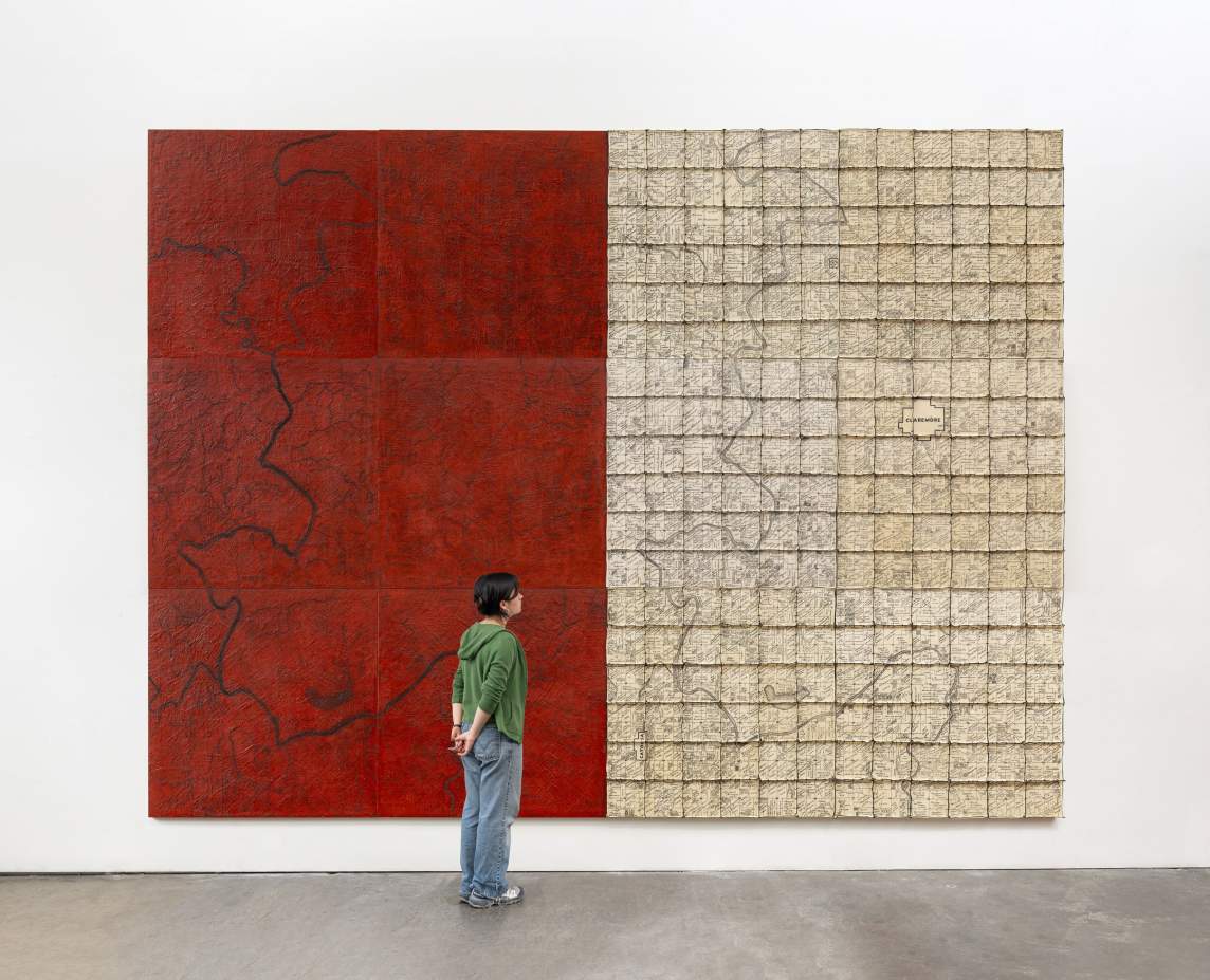 Old Homeplace is a large abstract diptych, more than twice as tall as the person standing in front of it. On left, is a textured red wax panel, with incised dark outlines of rivers and creeks. On the right, is a grid of white maps showing allotment lines in the Cooweescoowee District of Cherokee Nation, railroads, names of people, crudely sewn together with hog rings, overlaid with the same waterways.