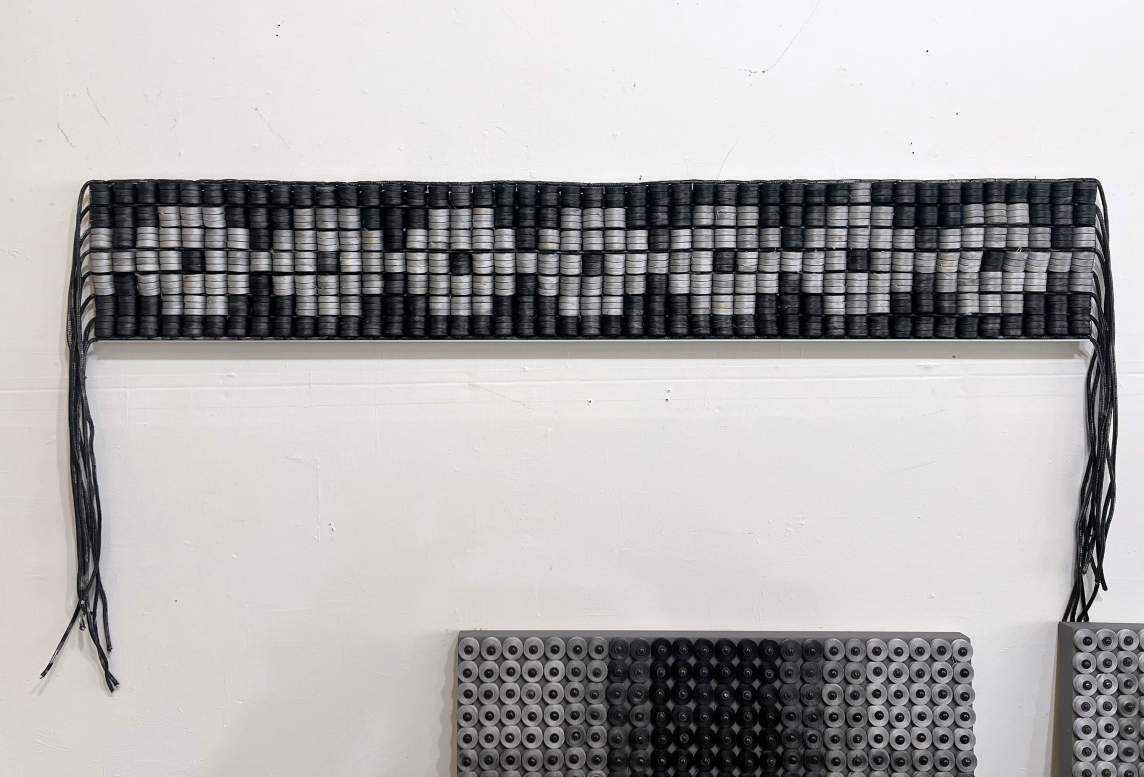 Forty-five columns of 21 black or silver spools are stacked together to make a long, woven strip, which sits on a shallow ledge against a white wall. The silver spools make angular O-shapes against the black. Thick cords hang down from the narrow ends, and racks of spools sit below the ledge.