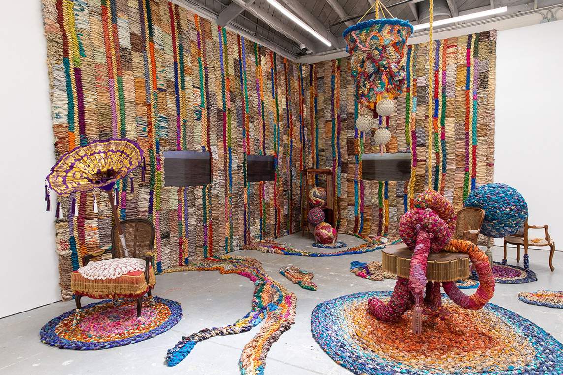 The corner of a gallery is hung with long floor-to-ceiling textiles made up of earth-toned and vividly colored fabrics. The hangings are woven with narrow or wider columns. Strips reach out from the bottom edge of the wall hangings in a few spots. Tables and chairs between the walls sit on round braided rugs and hold oversized cloth coils and balls.
