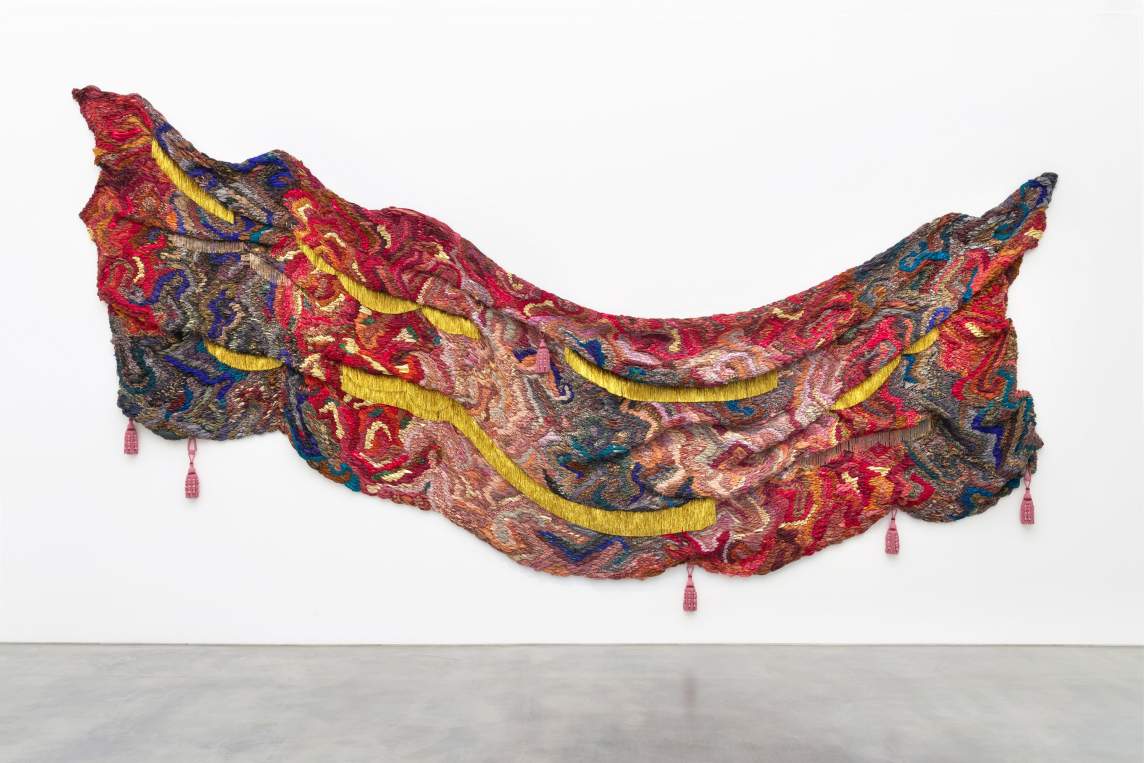 A rippling swath of a woven textile curves like a wide, shallow U across a white wall. The piece is mostly made with shades of red and pink with a few touches of blue, and canary-yellow lines cut through the otherwise marbled coloring. Five irregularly spaced pink tassels hang from the bottom edge, and a sixth tassel hangs from near the top edge.