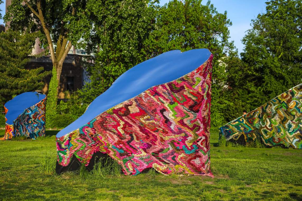 We are nomads, we are dreamers shows 3 abstract sculptures on a grassy field in front of trees. Each sculpture is an organic monolith-like form with the outer walls covered with sari fabric woven together in undulating wavy forms, bisected at an angle with a mirrored surface facing the sky.