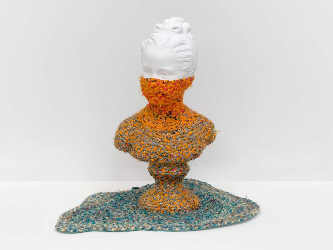 A white sculptural bust of a woman with her hair tied in a bow atop her head is nearly consumed by colorful braided coils, which cover the base up to just below the eyes. The woven textile on the bust is mostly marigold orange, and the puddle-shaped rug on which it sits is sky blue.
