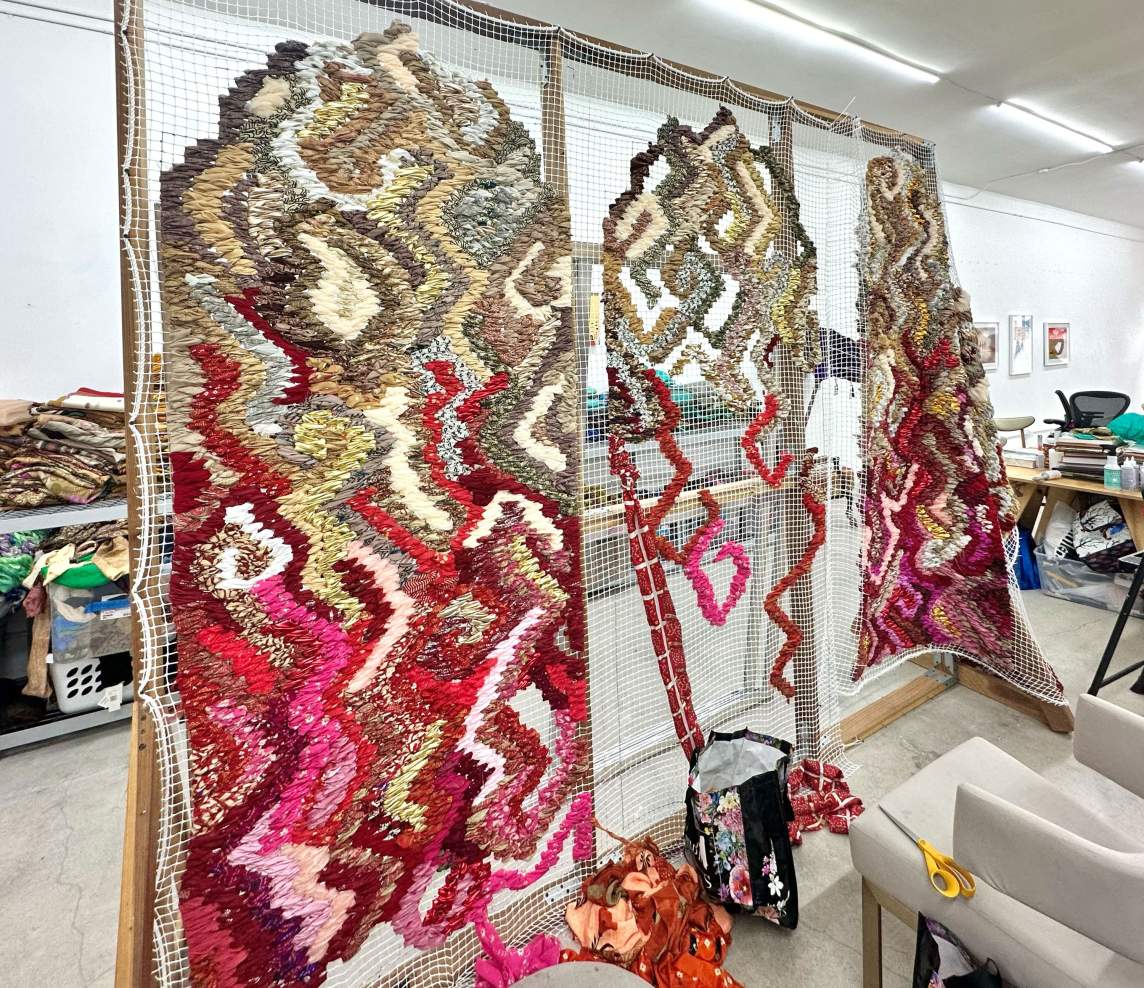 A grid of white mesh is stretched across a wooden frame in a studio. Strips of fabric in carnation pink, brick and garnet red, white, gold, and earthy tan are woven into three vertical sections.