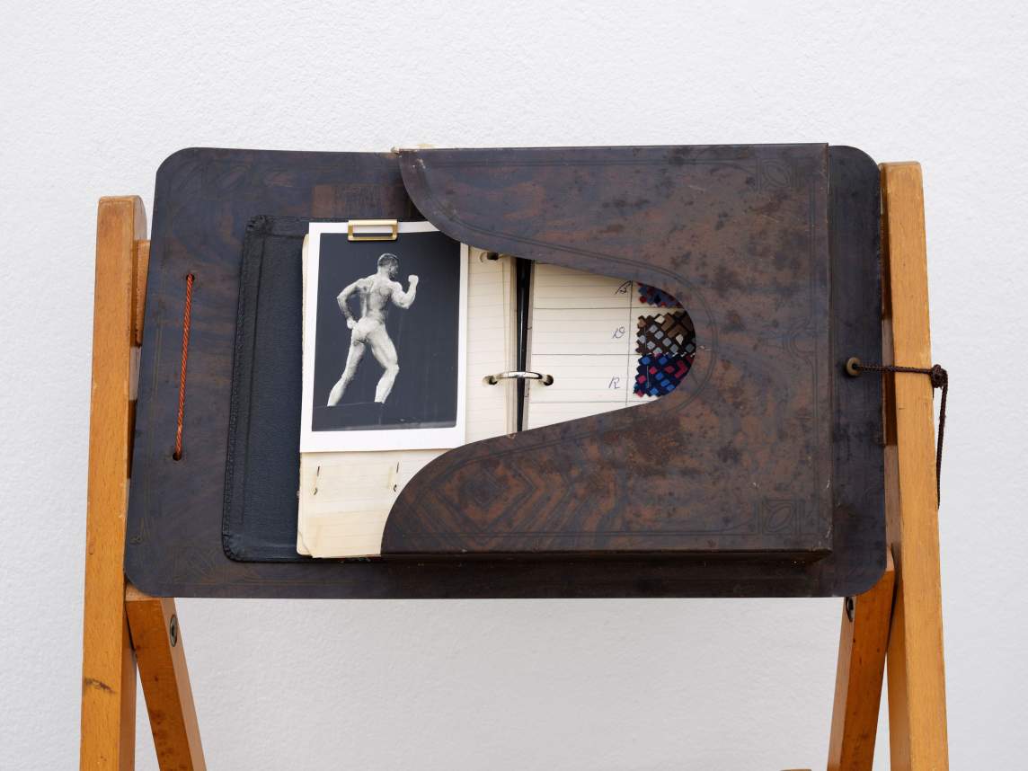 A document carrier like a thick portfolio is set so the opening is to our left. Half-visible inside is an open three-ring binder. A black-and-white photograph of a body builder is clipped to the left page, and scraps of fabric are affixed to lined paper to our right.