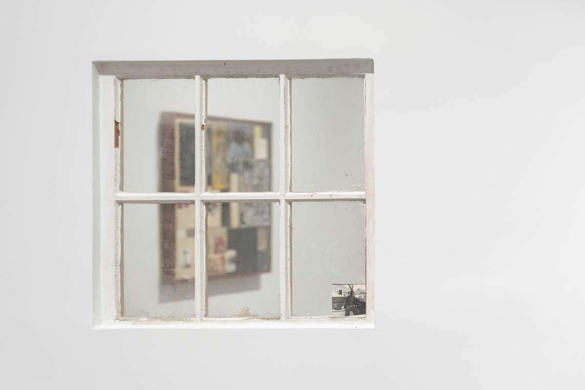 A scuffed, six-paned window is set into a white gallery wall. A black-and-white photograph of a Black man is set in the lower right corner of the window. The collage seen through the glass is softly out of focus.