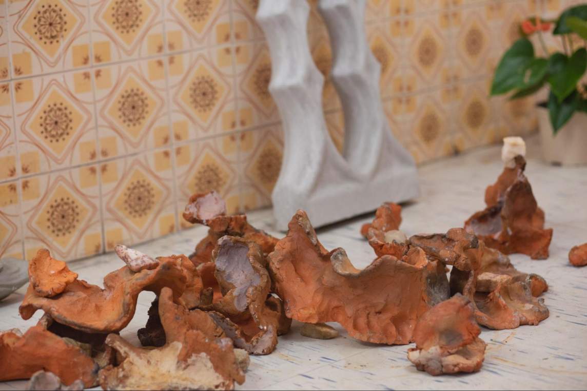 In a close-up detail of an artist’s architectural installation, a decorative cast concrete block leans against a patterned wall, amidst shards of red earthenware pottery, formed into organic shapes. A potted plant sits in the back right corner.