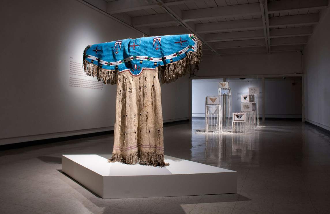 In a darkened museum gallery with gray floors, a traditional Lakota dress is displayed with dramatic gallery lighting opposite a group of sculptural box-like forms with dangling white threads and geometric imagery.