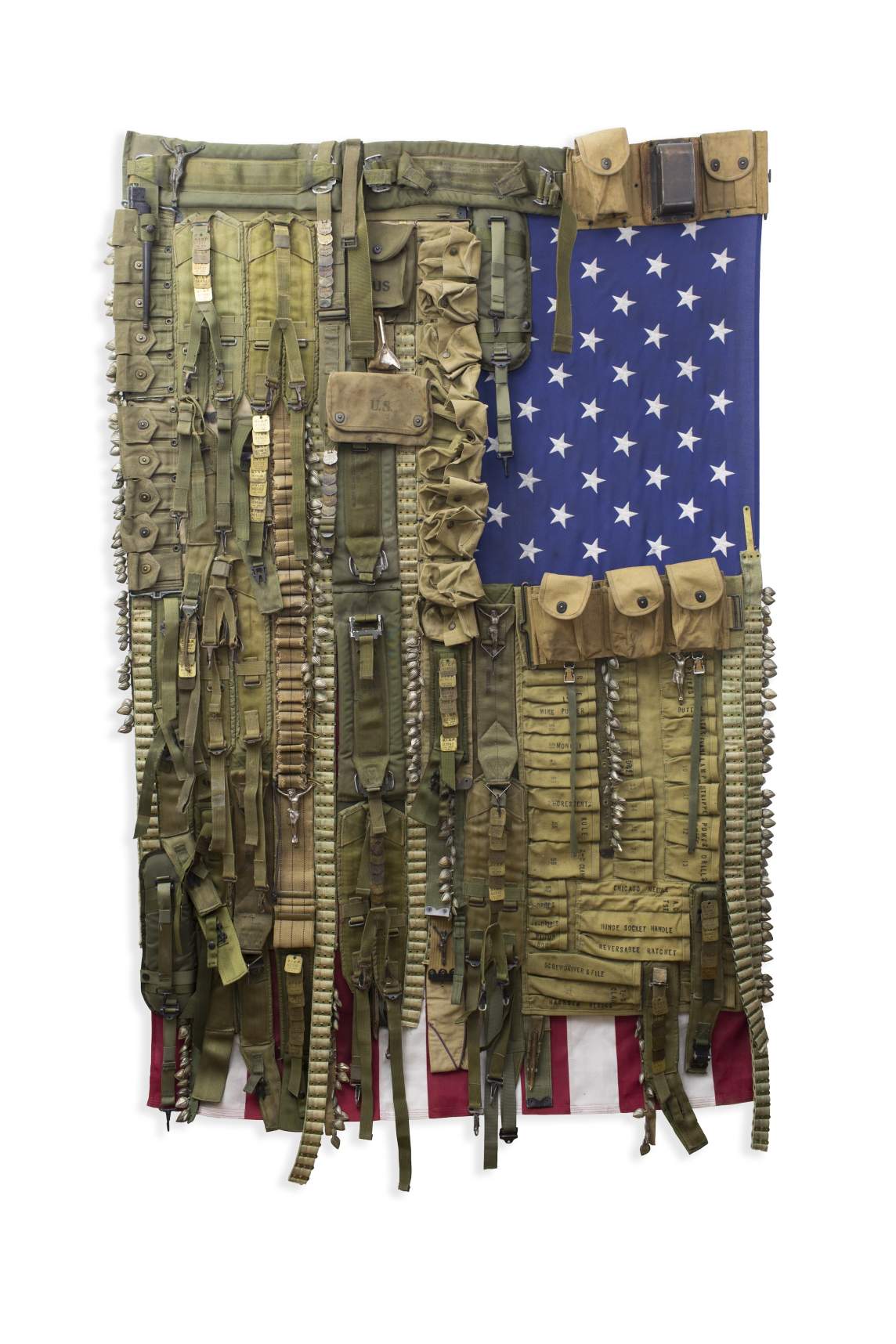 A US flag hangs facing downward, the stars at the top right corner. Orderly rows of military-style pouches, belts, and straps in fatigue tan and green entirely cover the stripes except for a strip along the bottom of the flag.