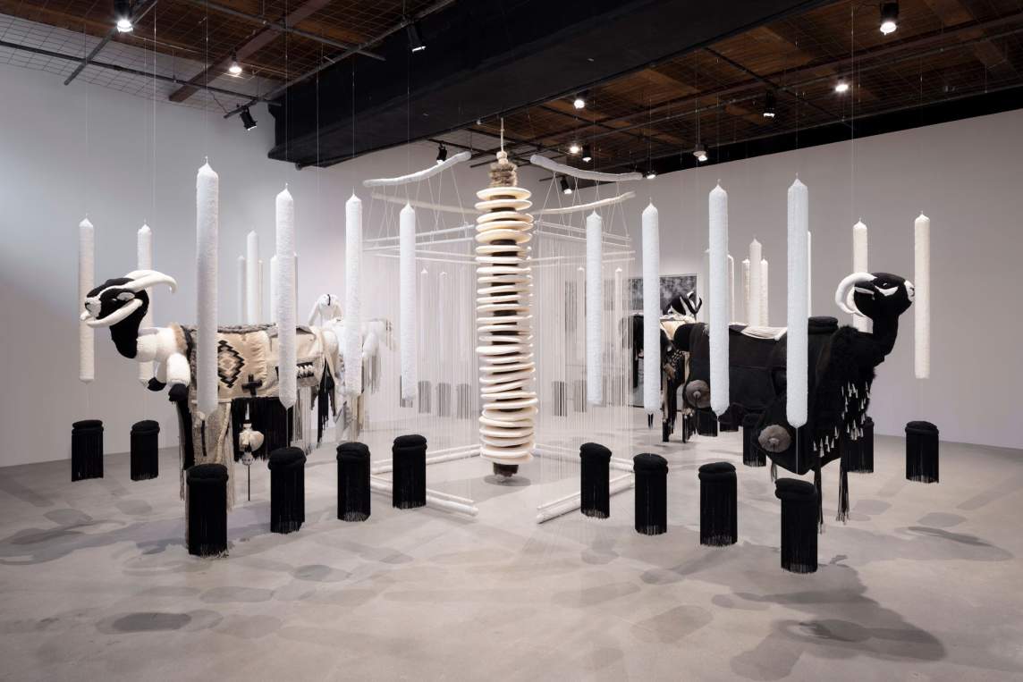 + [pronounced t’] is a sculptural installation of black and white components suspended from the ceiling featuring stylized ram sculptures, rows of tall cylindrical white candle-like forms above black cylinders, extending in iterations from a central vertical structure made of stacked disks.