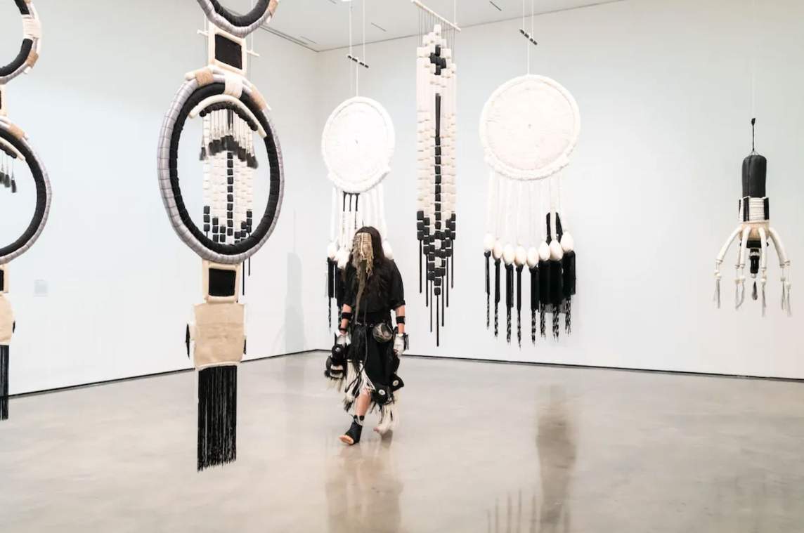 The artist walks through a gallery hung with six sculptures in tones of white, black, and tan.Four pieces have central woven hoops with fringe and weavings hanging down. Another appears to be woven into a vertical banner, and the fourth has a columnar body with eight white arms or legs curving out from near the bottom.