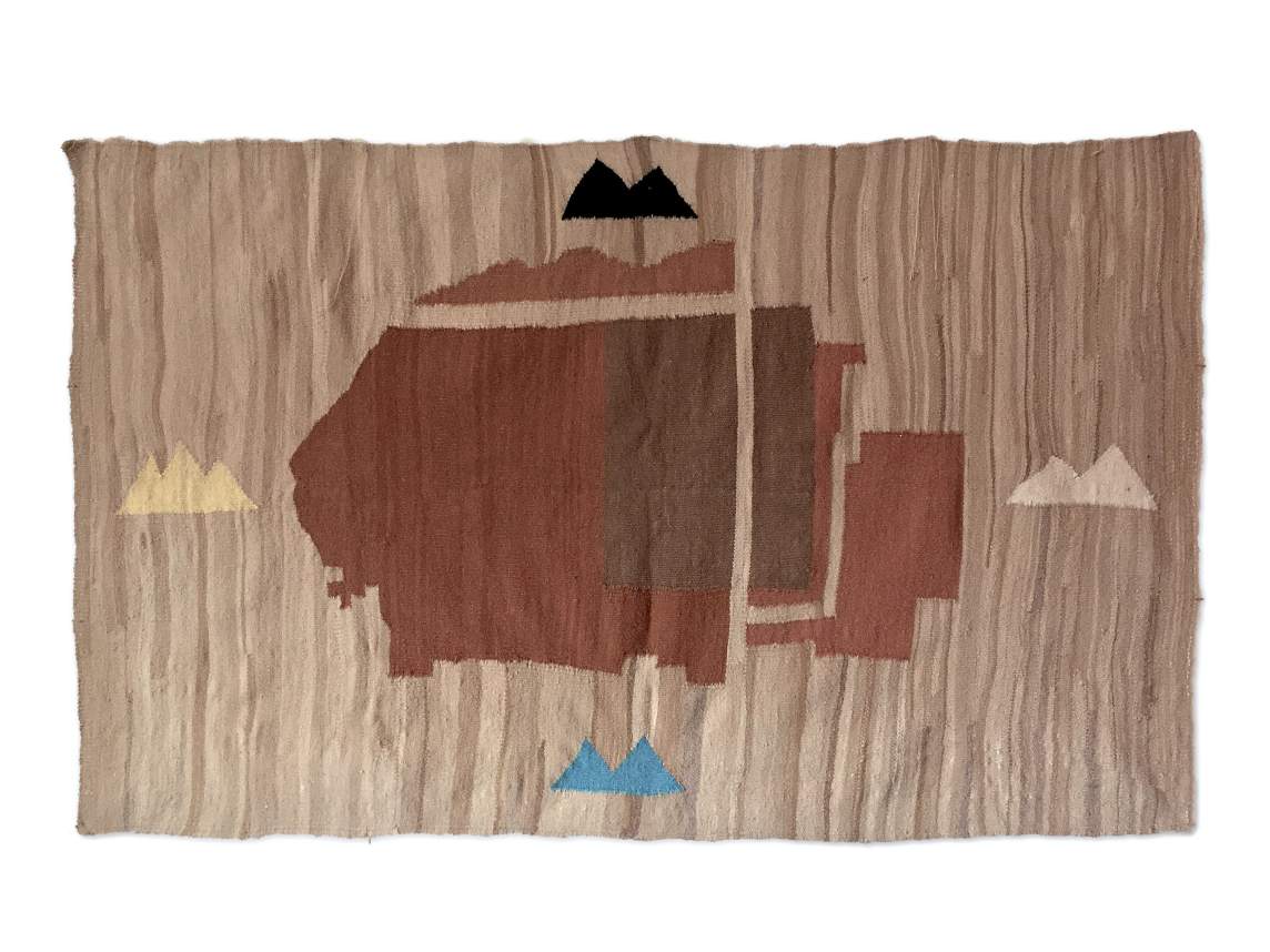 A rectangular flag is woven with a brown form at the center, possibly an animal, against a tan-colored background. To each side and much smaller in scale are double or triple-peaked mountains with black above, white to the right, blue below, and yellow to the left.