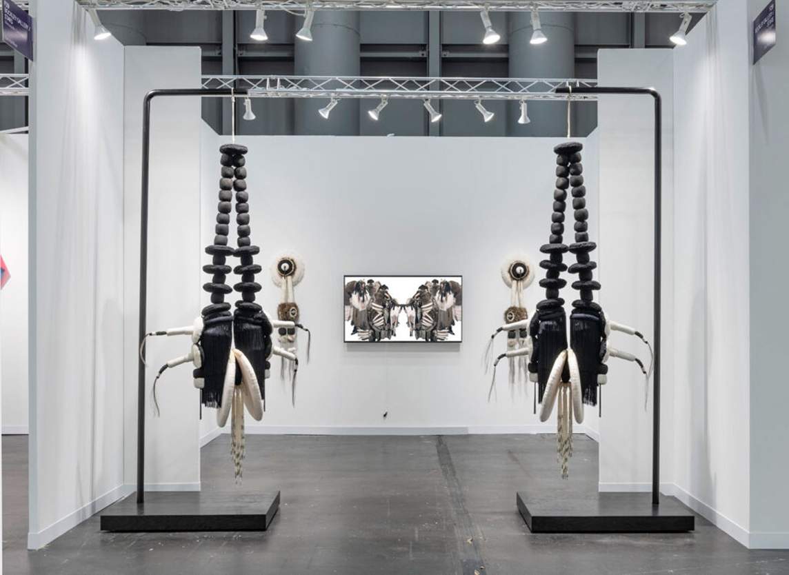 Installation view of two black and white sculptures hanging from metal frames. Each sculpture is made up of oversized black beads leading down to black, shell-like shapes and white rings that all together resemble a downward-facing fish. Two sculptures and a framed work hang on the wall beyond.
