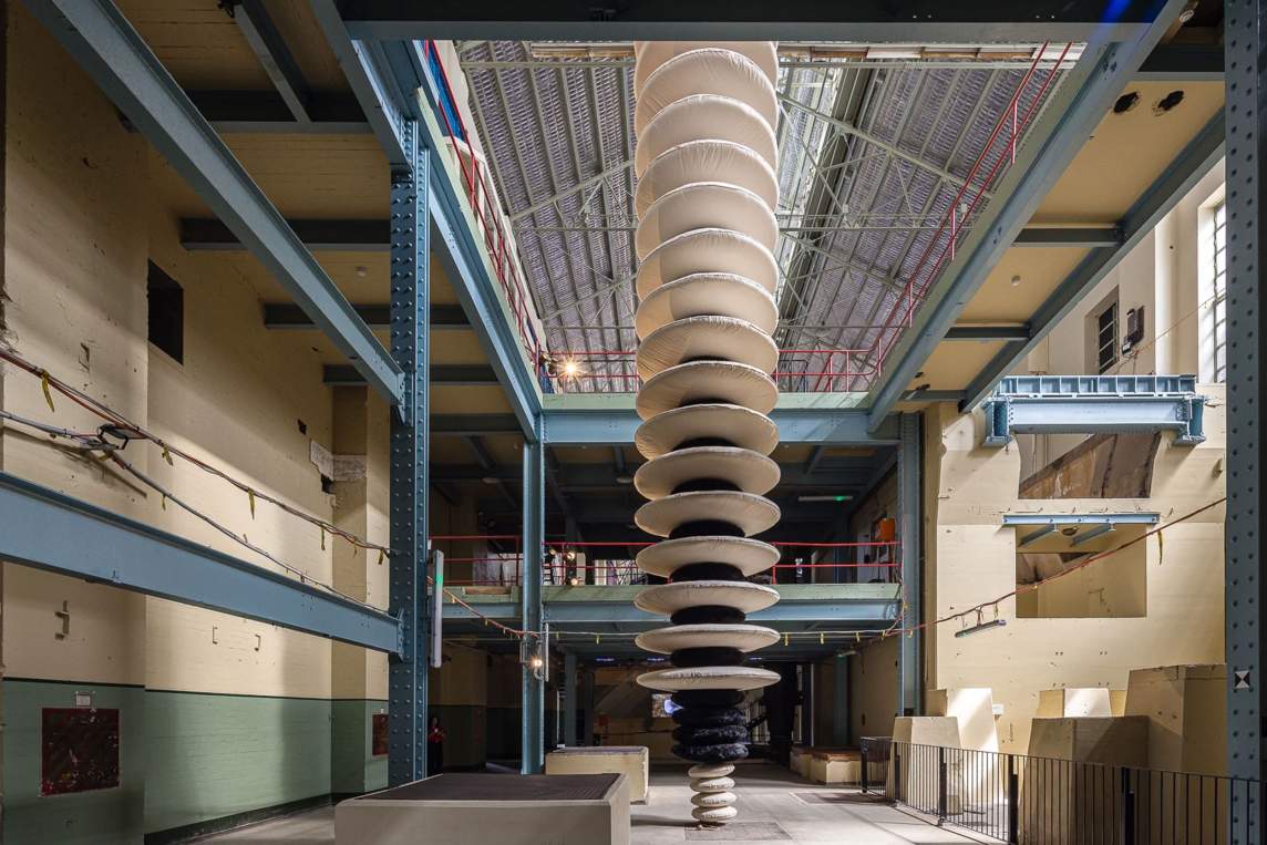 A column of disks reaches from the floor of an atrium in an industrial building, up two stories, and then disappears into a peaked roof above. Four white disks, the smallest, are at the bottom of the column. Four slightly wider black disks follow before the black disks alternate with the largest white disks all the way up the column.