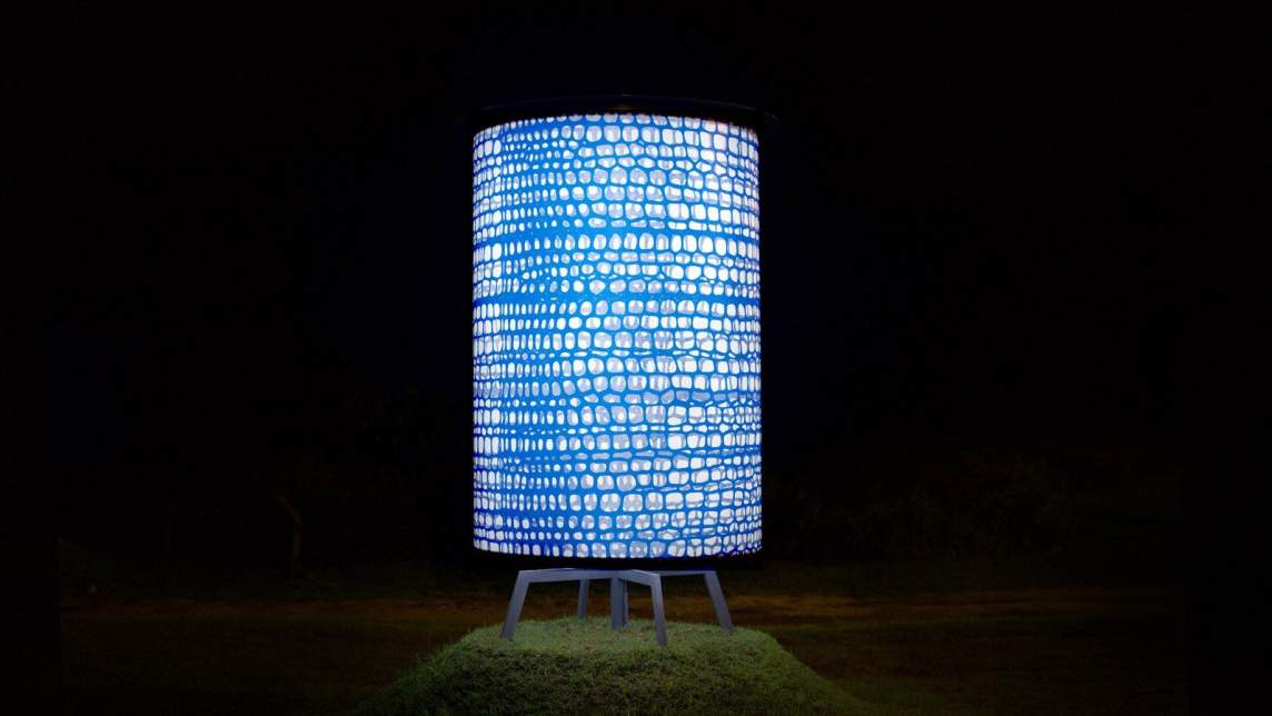 At night, a large metal cylinder illuminated from within stands on a small grass hill. The cylinder is covered with a print of blue webbing pattern.