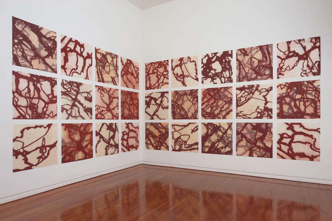 A series of 24 woodcut prints, each with a different pattern of meandering iron red lines, are installed in a grid across two walls an art gallery with white walls and shiny wood floors.