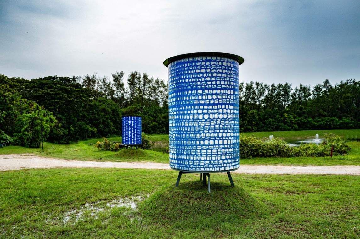 On a field of green grass with a tree line behind it, two large metal cylinders are installed in an art installation. Each is covered with a print of blue webbing pattern.