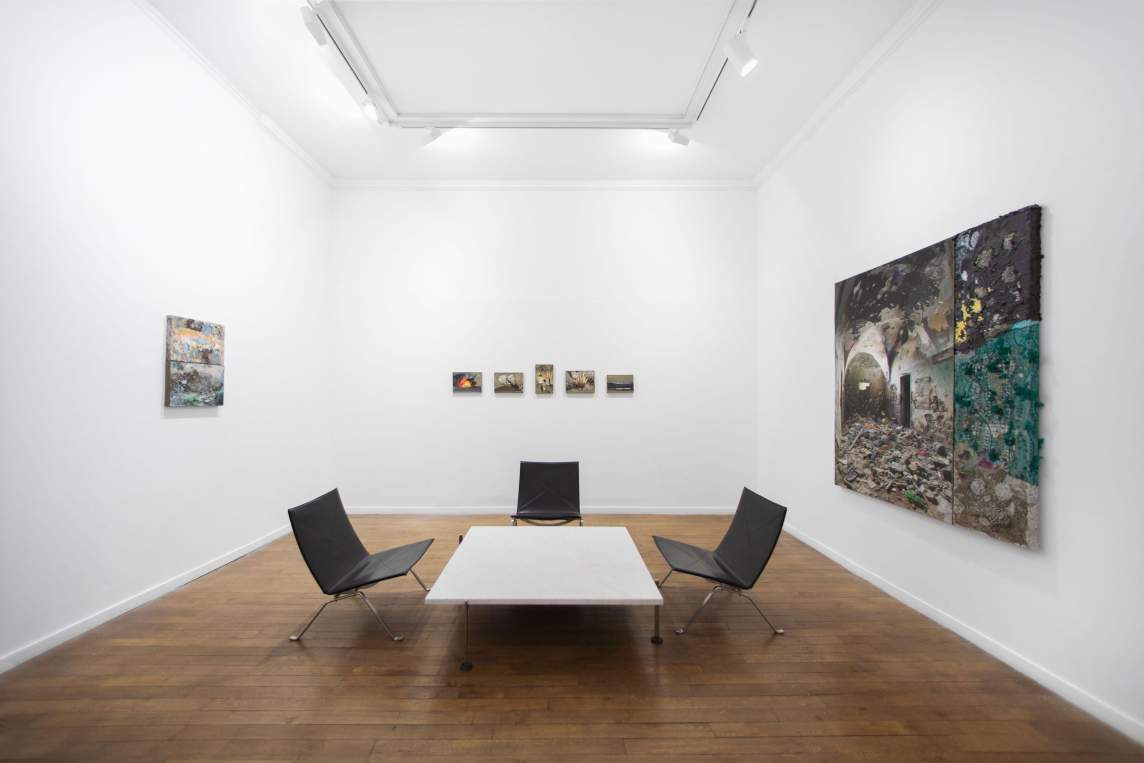 In an art gallery with white walls and a wooden floor, artworks of various sizes hang on three visible walls. In the center of the room is a low white table surrounded by four black chairs.