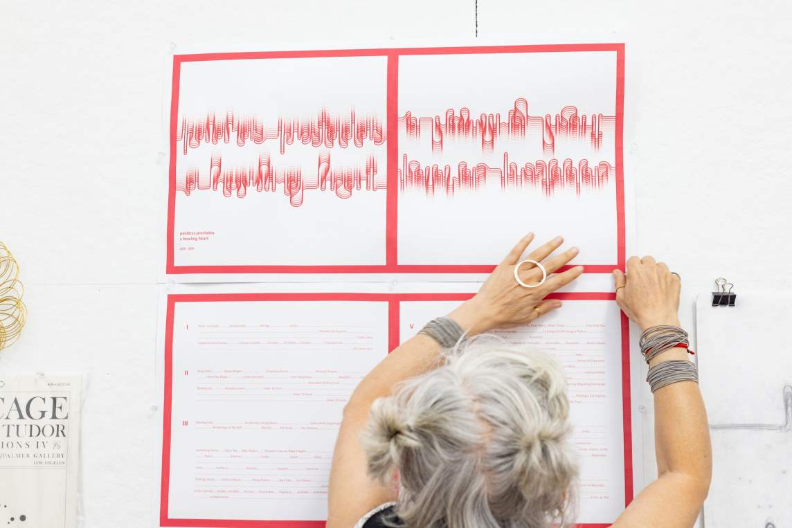 Analia Segal reaches up to tack artwork to the wall. The work has an abstracted word form running across in red, surrounded by a graphic red frame