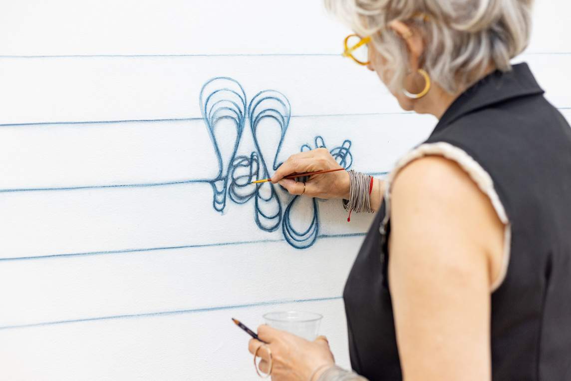 Analia Segal paints with blue watercolor a looping abstracted word form directly on a white studio wall. She is an Argentine-American artist with grey hair pulled up into two small ponytails, light skin tone, and wears a black vest, lined pants, and silver chain bracelets.