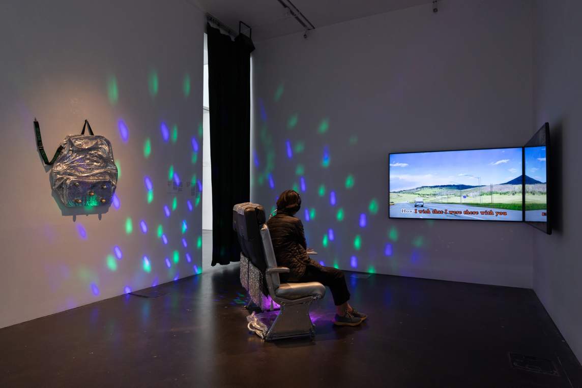 Two airplane seats sit in a gallery in front of a pair of large-screen TVs. On the visible screen are the words “I wish that I were there with you” overlaid on a landscape. Purple and green lights projected from near the seats create dashes on two of the walls in the otherwise dim gallery. A silver piece of luggage hangs on the wall opposite the screens.
