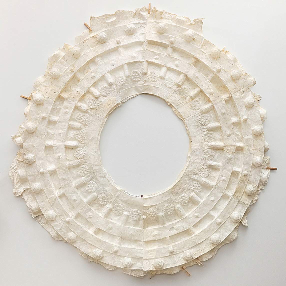 Cream-white paper has been pressed to create a wide ring with an open center. The form is made up of concentric rings, of rows of bosses like little mounds, and a ring of gears. Clothespins hold the irregular, ruffled edges on the white wall.