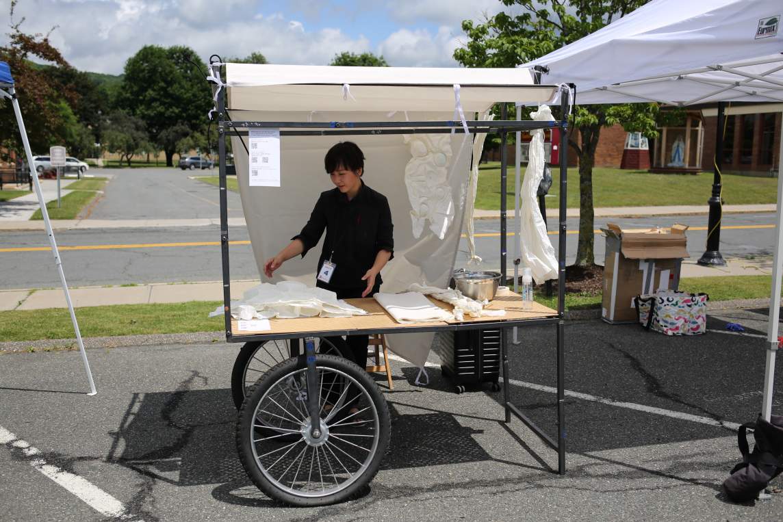 The artist stands behind a cart held up on two oversized, bicycle-style wheels and a metal frame holding a wood top. A white cloth canopy creates shade on a sunny day. White material and a silver bowl are laid out on the table.