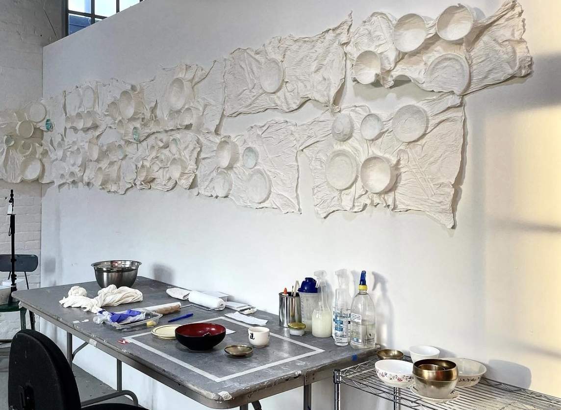 At least 15 white sculptural pieces are hung closely together in two rows on a temporary wall behind a work table. On each piece, the white, wrinkled material presses closely around bowls, dishes, and silverware.