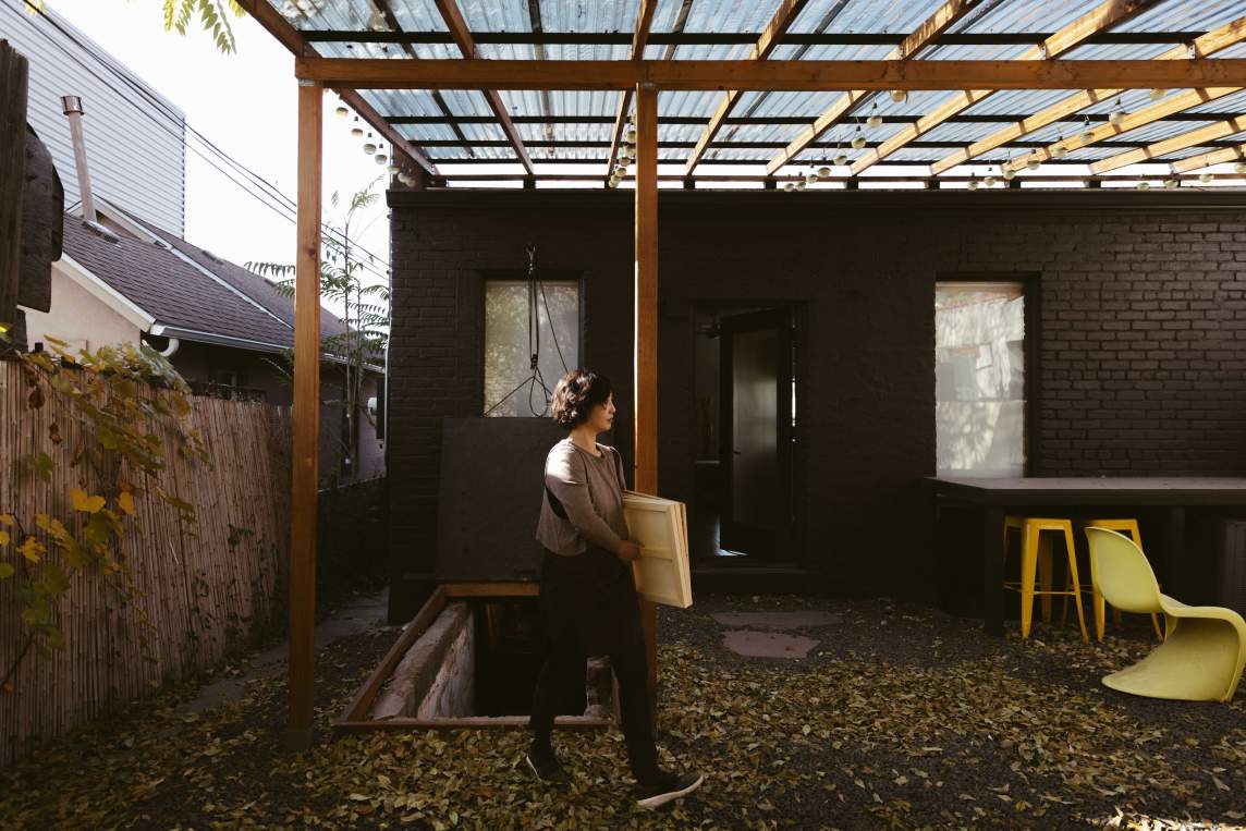 The artist carries three wood panels tucked under one arm in a back yard. Under an awning at the back of a house, she walks past an opening leading to underground stairs.
