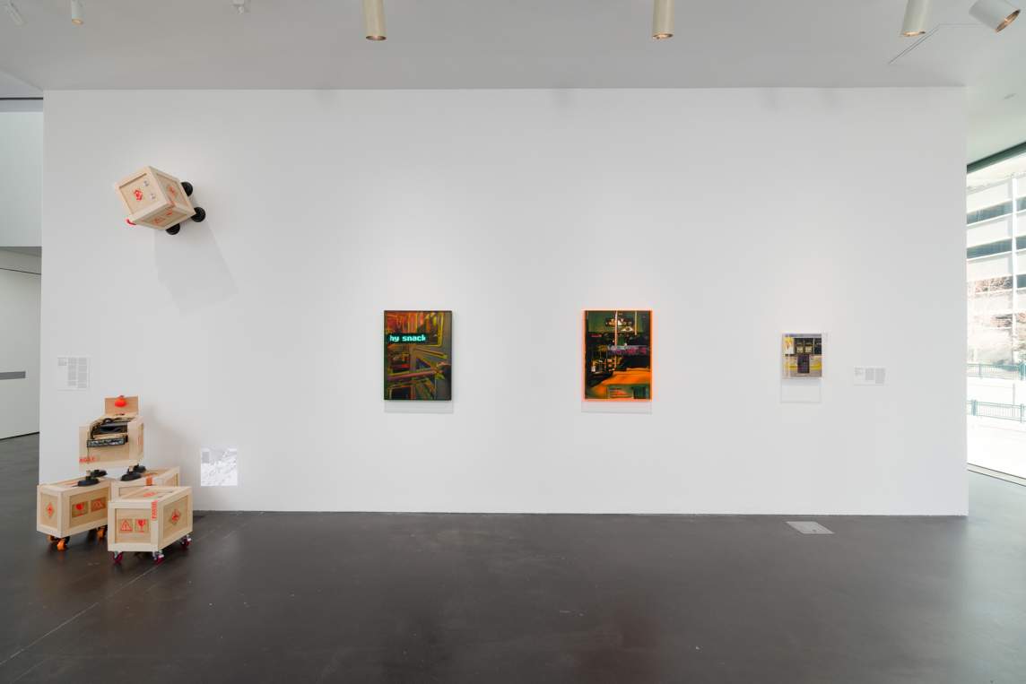 Installation view of three works hanging on a white, floating gallery wall. To the left are five wooden crates. All are made with pale wood painted with vivid orange warning symbols. Three resting on the floor are supported on wheels. The other two have plungers for feet. One of that pair perches on two of the crates with its top open. The fifth is stuck to the wall by its plunger feet high above the others.