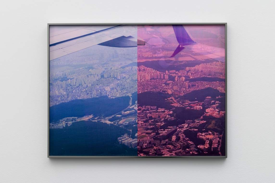 An aerial view from an airplane window down onto a city. The photograph is split vertically into two halves. Both are monochromatic like a black-and-white photograph, but the left half is shades of violet and ashy purple, and the right half is saturated raspberry pink. The two halves almost, but do not totally, line up.