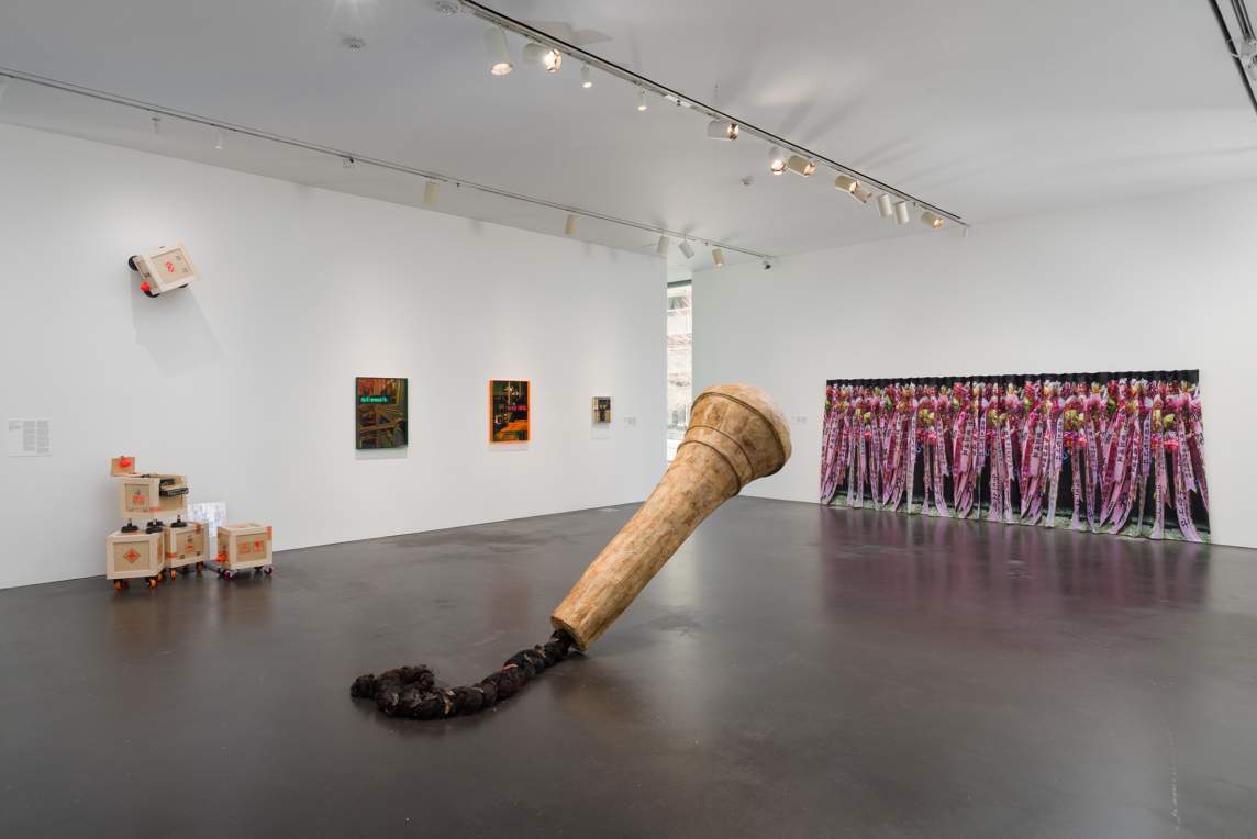 An oversized microphone in a gallery installation appears to be carved from wood. The microphone hovers over the floor at a 45-degree angle, and a thick black rope coils off its back end. Works hang on the walls opposite us, and a sculpture made up of five wooden crates is to our left.
