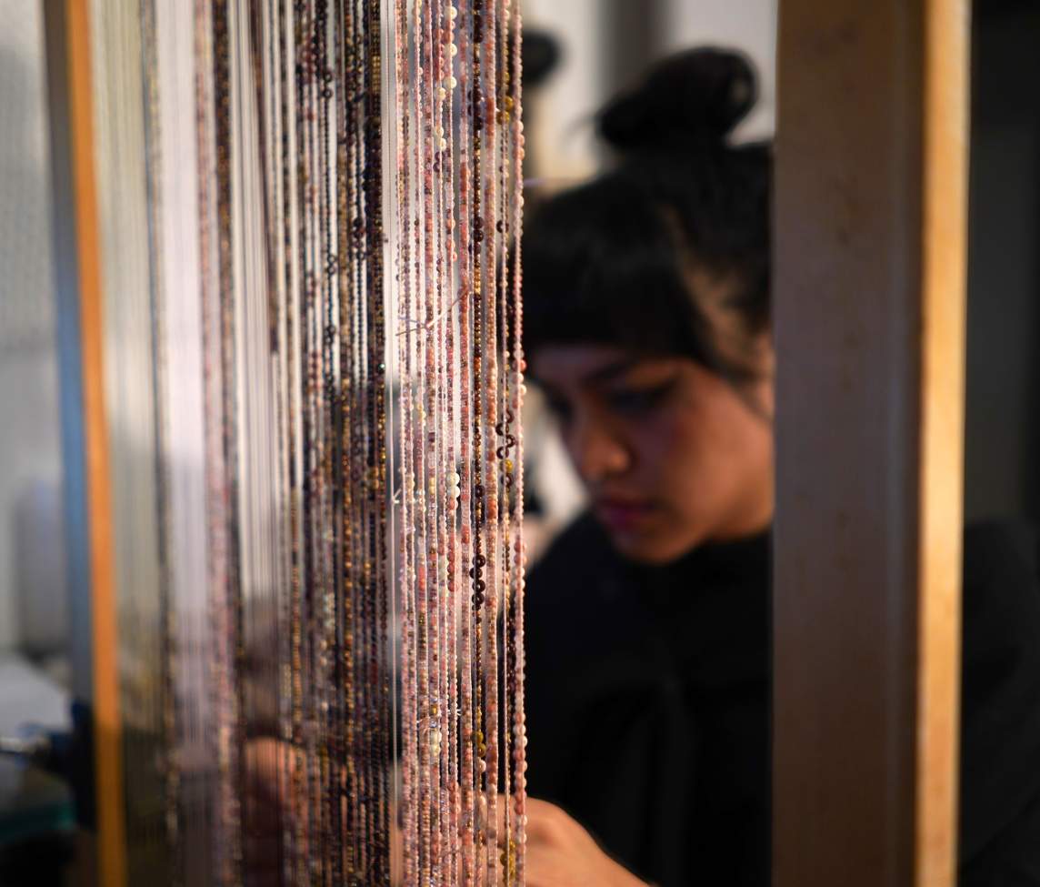 Threads strung vertically with pink, black, and brown beads are spaced tightly across the center of a wood frame. Out of focus, the artist looks down at her hands in the background.