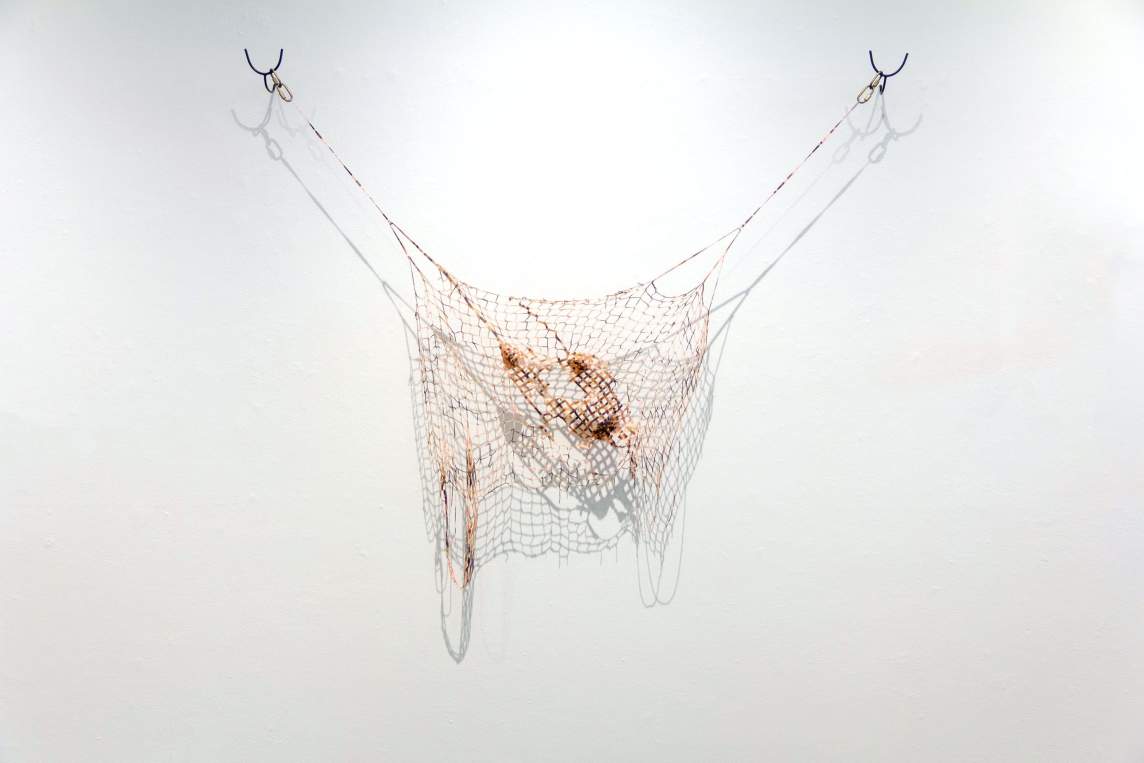 Loosely woven mesh in shades of brown hangs limply from two long cords that stretch to either side from the top corners. The area around a hole in the weaving facing us darker brown. The hanging cords end in pairs of snap hooks, which are looped over U-shaped brackets screwed into the wall.