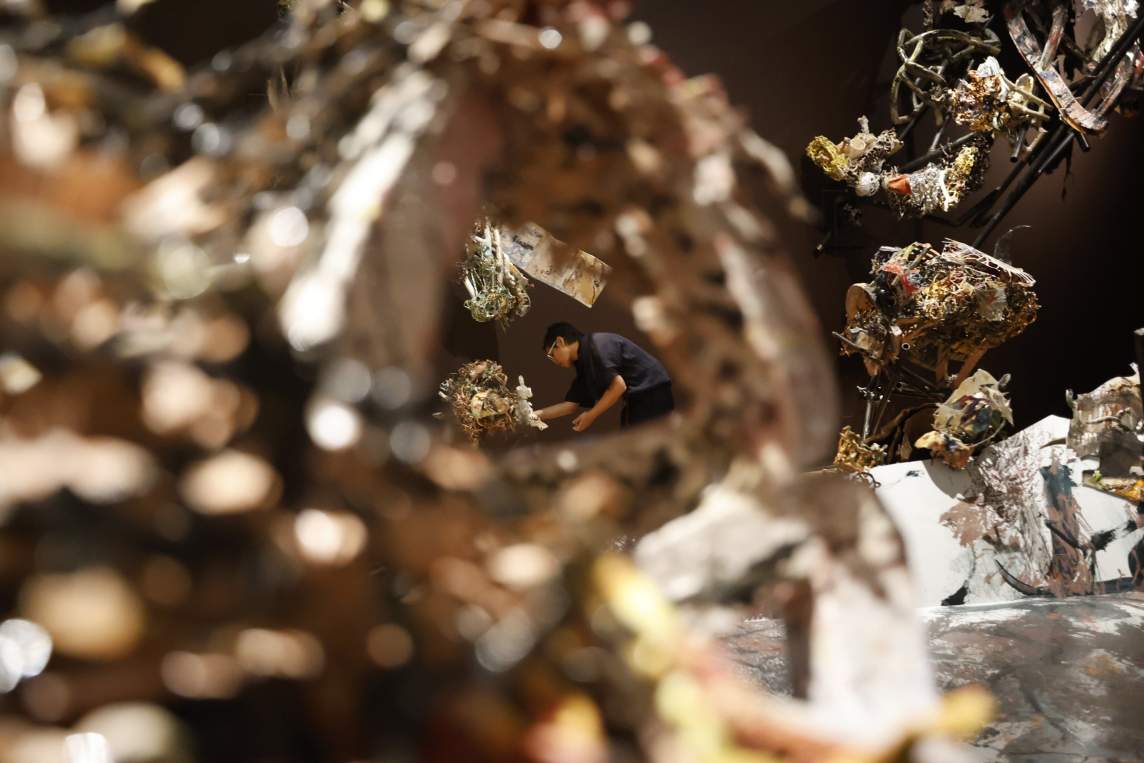 Seen through an opening in an out-of-focus abstract ceramic sculpture, Linda Sormin bends down to adjust an abstract sculpture of ceramic tangles. She wears dark clothing and glasses, and has short dark hair.