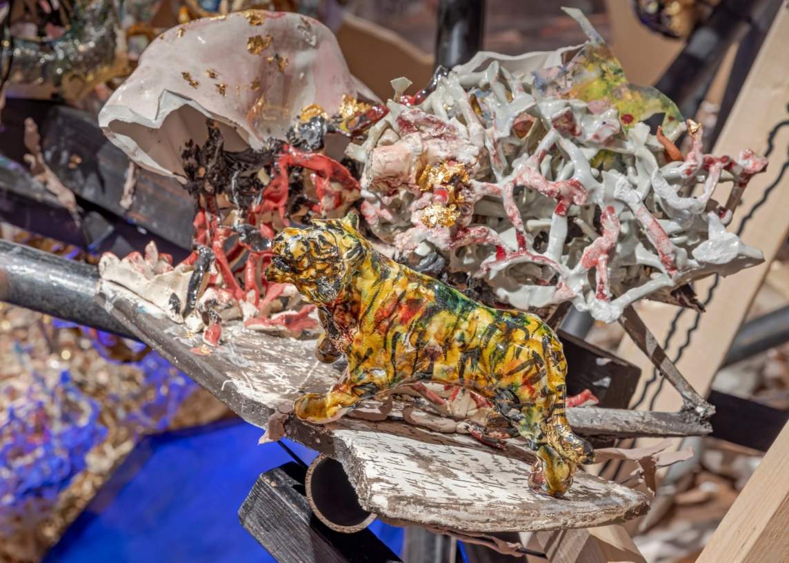 The green- and black-streaked yellow figurine of a tiger stands in front of ceramic shards and basket-like forms. To the right, behind the tiger, white strips are stained red or covered in gold leaf in some areas. To the left, a solid piece of ceramic drapes like a jellyfish above jagged, broken edges.