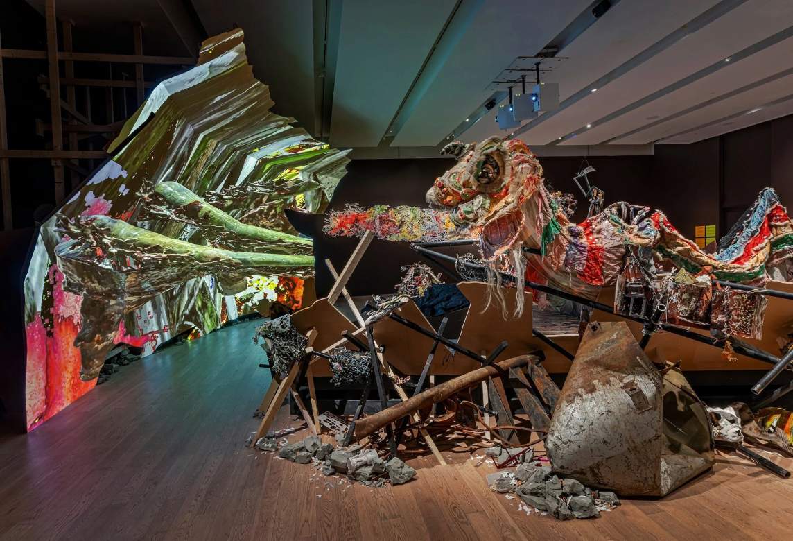 The tattered form of a Chinese dragon costume vies for attention with a jumble of metal or wood poles, sticks, rocks, bent sheet metal, and other debris on a hardwood floor in a gallery space. Green arms to the left seem to reach out to an upward-curving backdrop streaked with mossy and pine green with a few hits of orange.