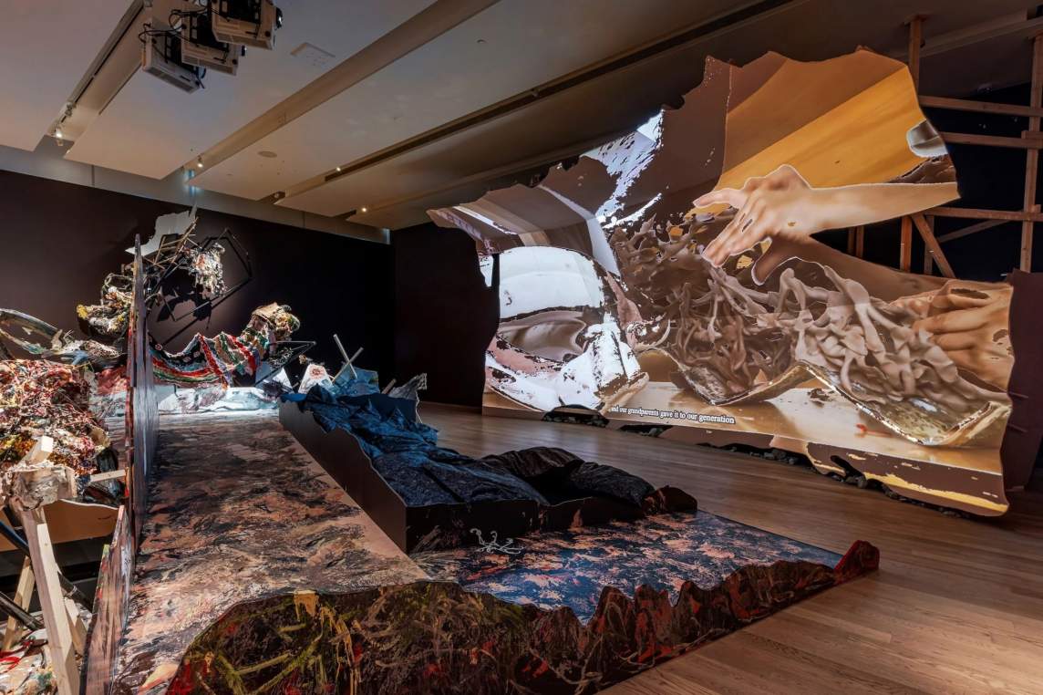 The dragon costume is at the far end of the room in this second view of the gallery space. Between the dragon and us is an expanse of molded material painted with streaks and slashes of earthy green, brown, red, and black. An image of tan and beige-colored forms is now projected on the curved screen to the side.