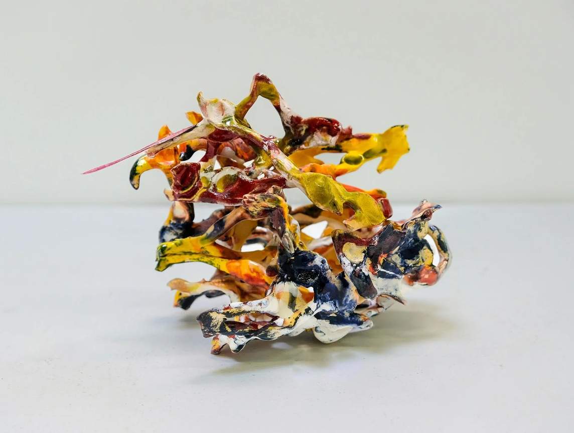 A roughly orb-shaped ceramic sculpture appears to be made from strips of canary yellow, burgundy red, midnight blue, flame orange, and snow white. The colors streak irregularly across the uneven surfaces.