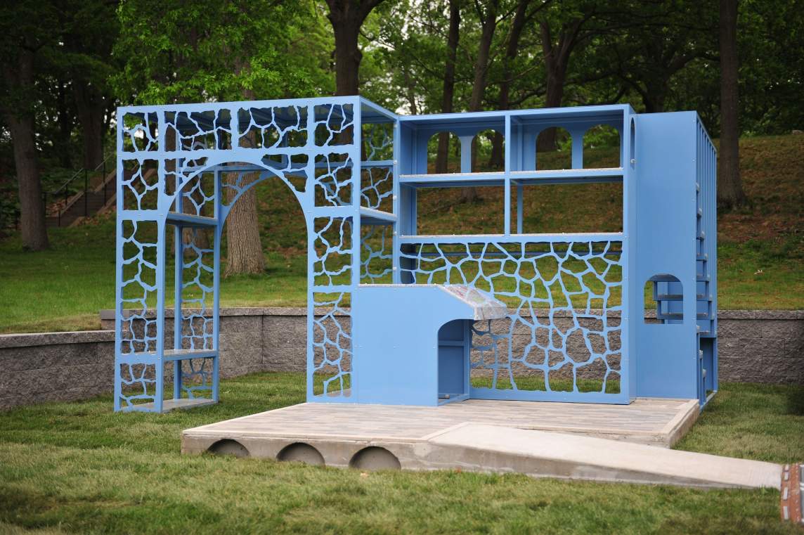 Matters of Consequence is a public sculpture composed of architectural details including a wall with shelves, small stairs, a desk-like shape, and shaped cutouts resembling the grout of stone work, flanked by an arch with cutouts, on a concrete stage with a ramp, and grass and trees in the background.