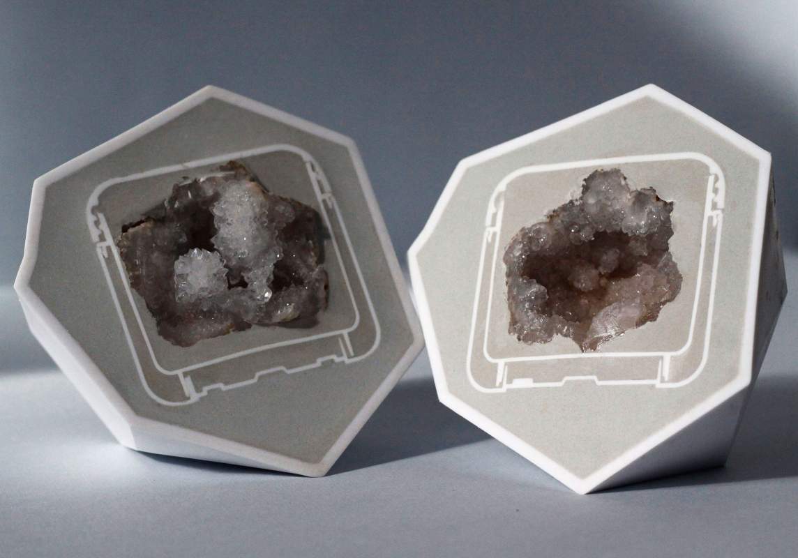 Two halves of an angular, seven-sided orb are splayed open to show silvery gray and frost white crystals at their centers. Each half has a white plastic shell with a concrete-colored face where a geode would have been split. The openings in each half are square and framed by an outline resembling a microchip.
