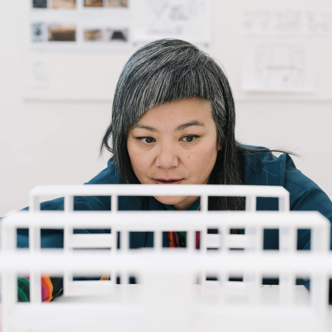 The artist looks across the top of a sculptural piece made up of three upright, white, free-standing panels. Drawings and papers hung on the wall behind the artist are blurry in the background.